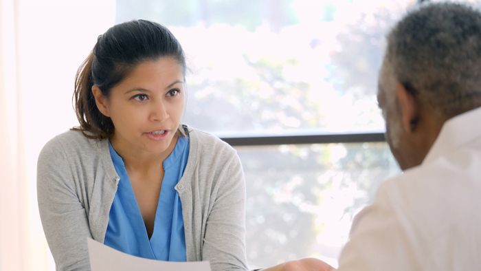 Close-up of female nurse sitting across from elderly male while consulting him with paper in her hand.