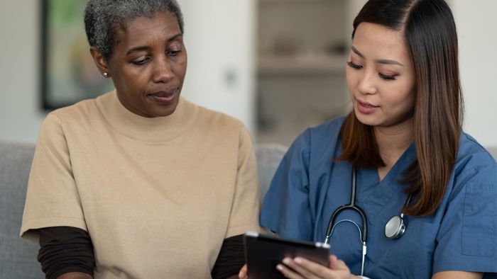 Close-up of female nurse and elderly female patient sat together looking at tablet in nurse's hand as she consults patient.